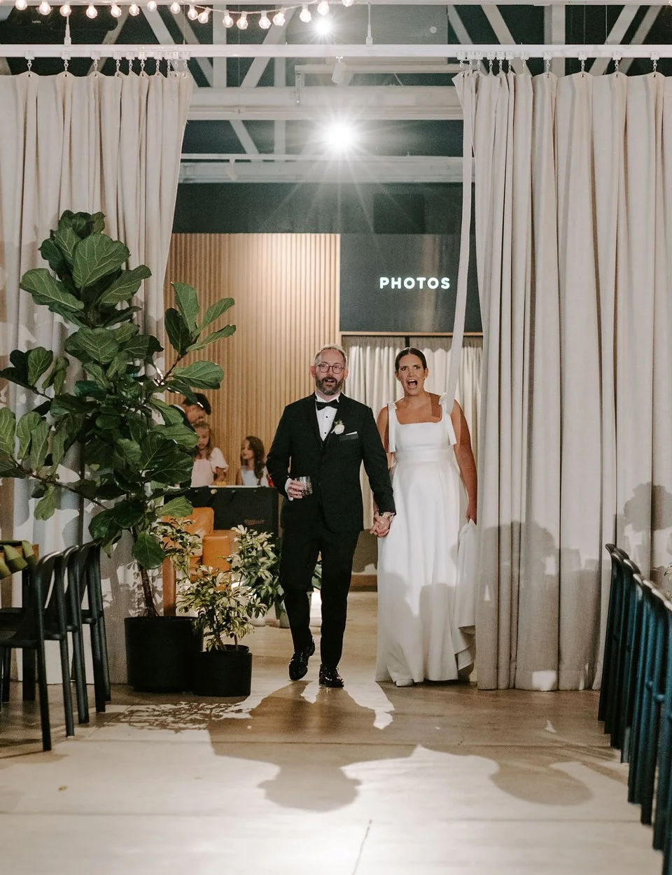 A newlywed couple, a man in a tuxedo and a woman in a wedding dress, holding hands and walking through a curtain in a wedding venue. The groom has glasses and a beard, and the bride has dark hair styled in an updo. In the background, there are some c