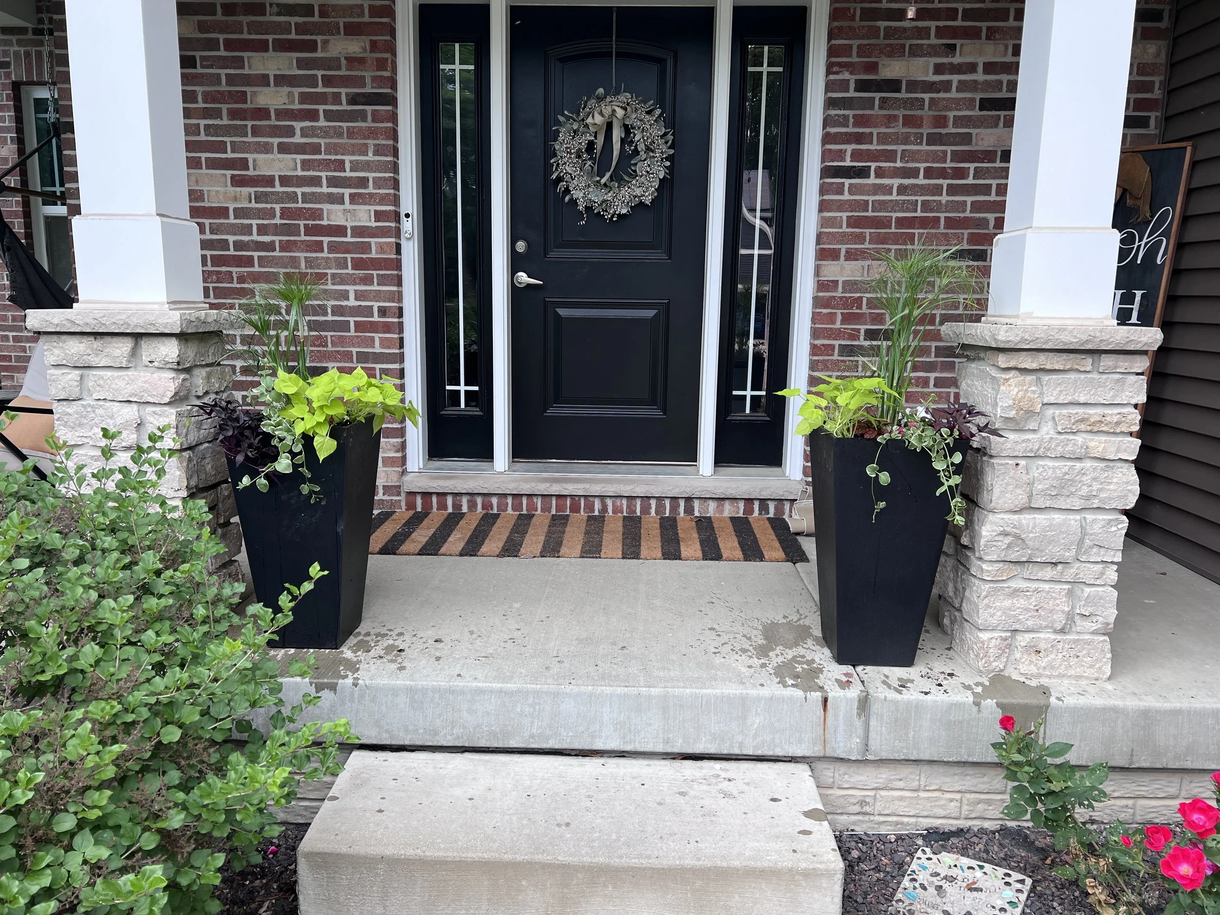 A front porch with a black door decorated with a festive wreath, flanked by two tall black planters with green plants, and a welcome mat in front of the door, with some flower pots on the side.
