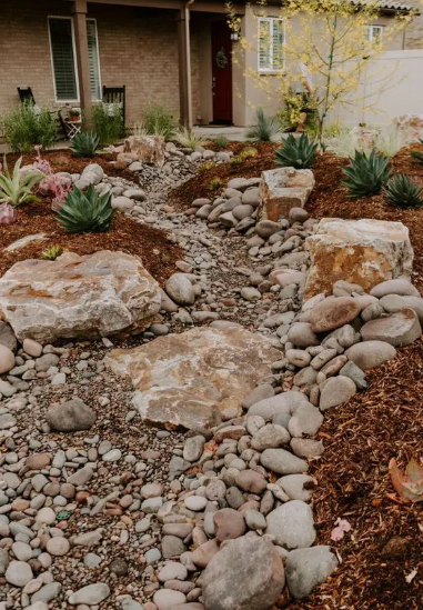 A landscaped front yard with a stone pathway, large decorative rocks, succulents, cacti, and mulch, leading to the house entrance.