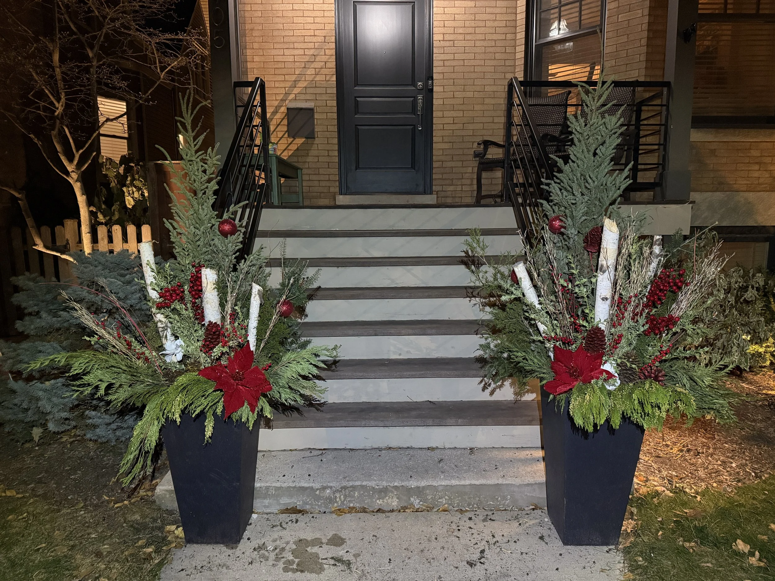 Decorative Christmas arrangements with red poinsettias, evergreen branches, white birch branches, red berries, and pine cones in large black planters flanking a front porch staircase with brick house in the background.