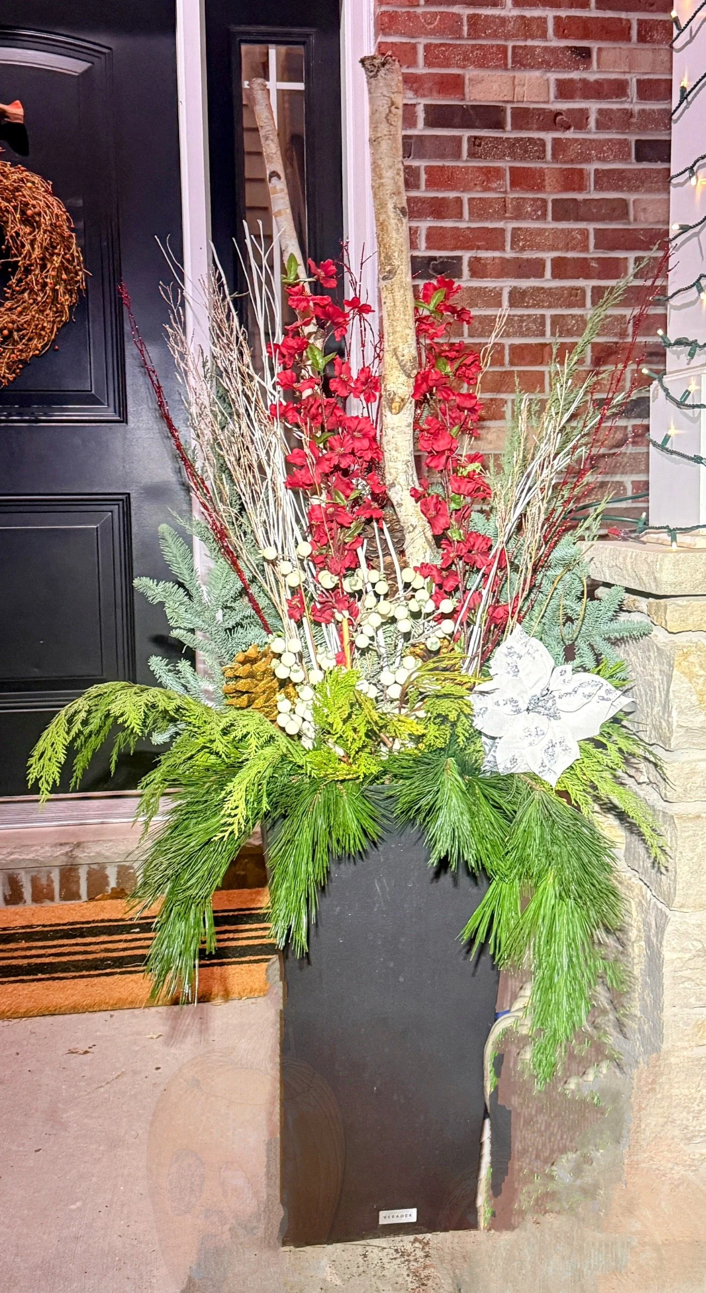 Festive holiday floral arrangement in a black vase, featuring red berries, pinecones, evergreen branches, and red flowers, placed outside a brick house front.
