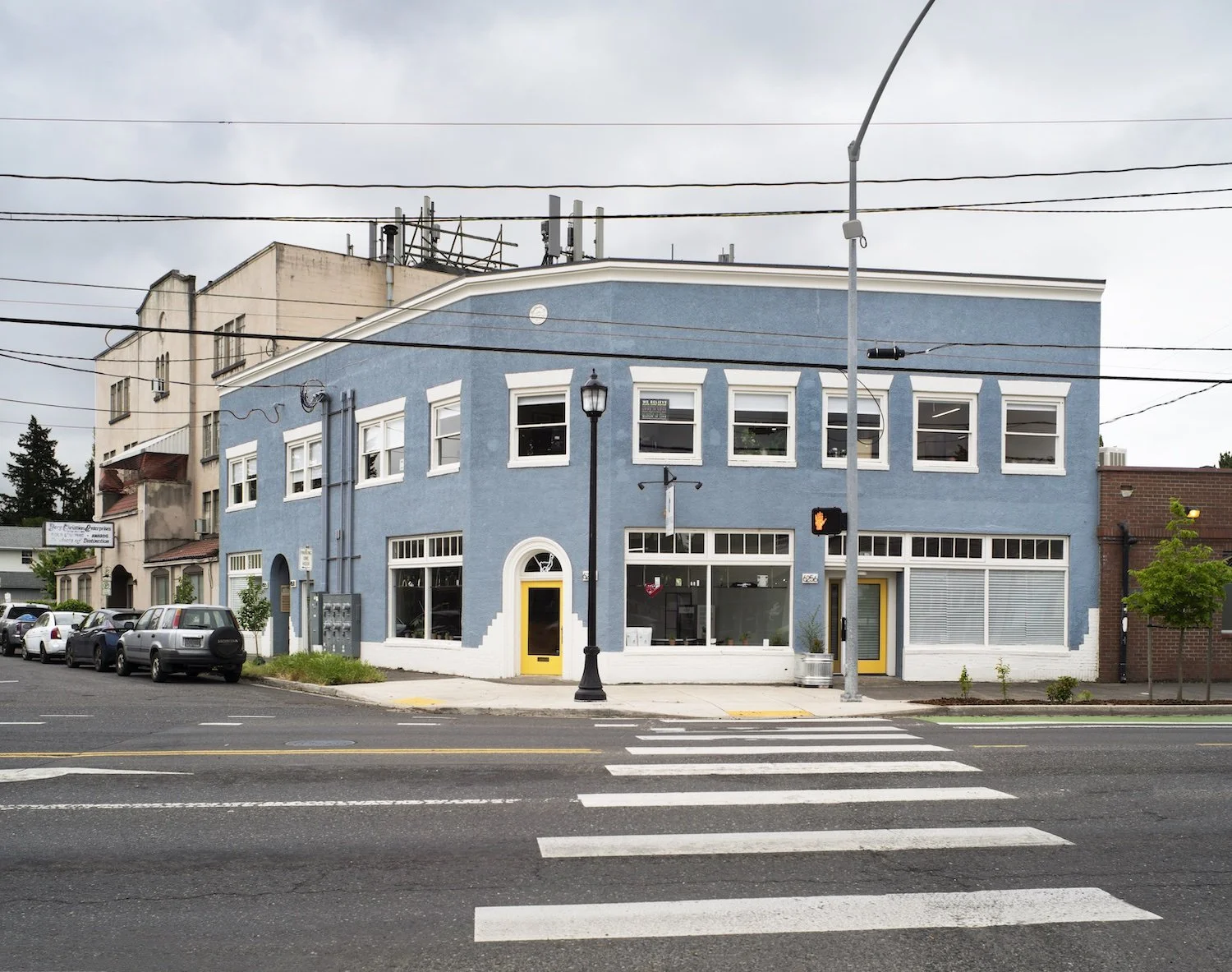 A two-story blue building with white trim and yellow door, located on a city street with cars parked in front, a crosswalk, and a traffic light.