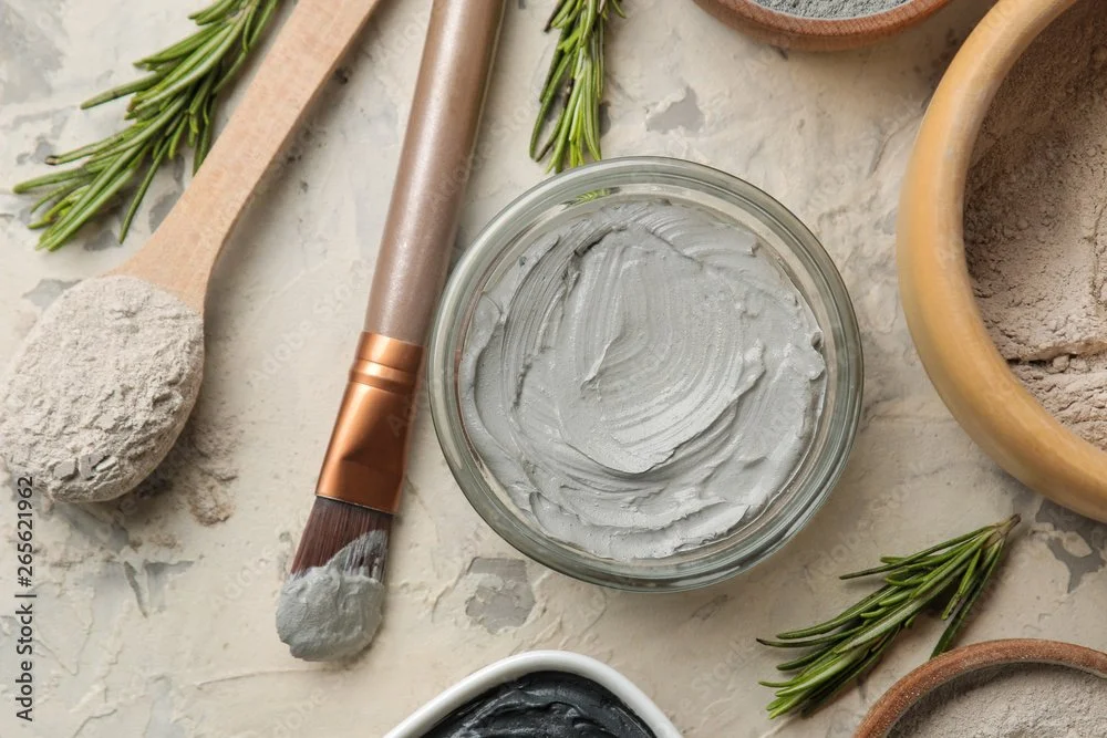 A glass jar of gray clay facial mask surrounded by herbal sprigs, a wooden massage roller with clay on its brush, and bowls of clay powders on a textured surface.
