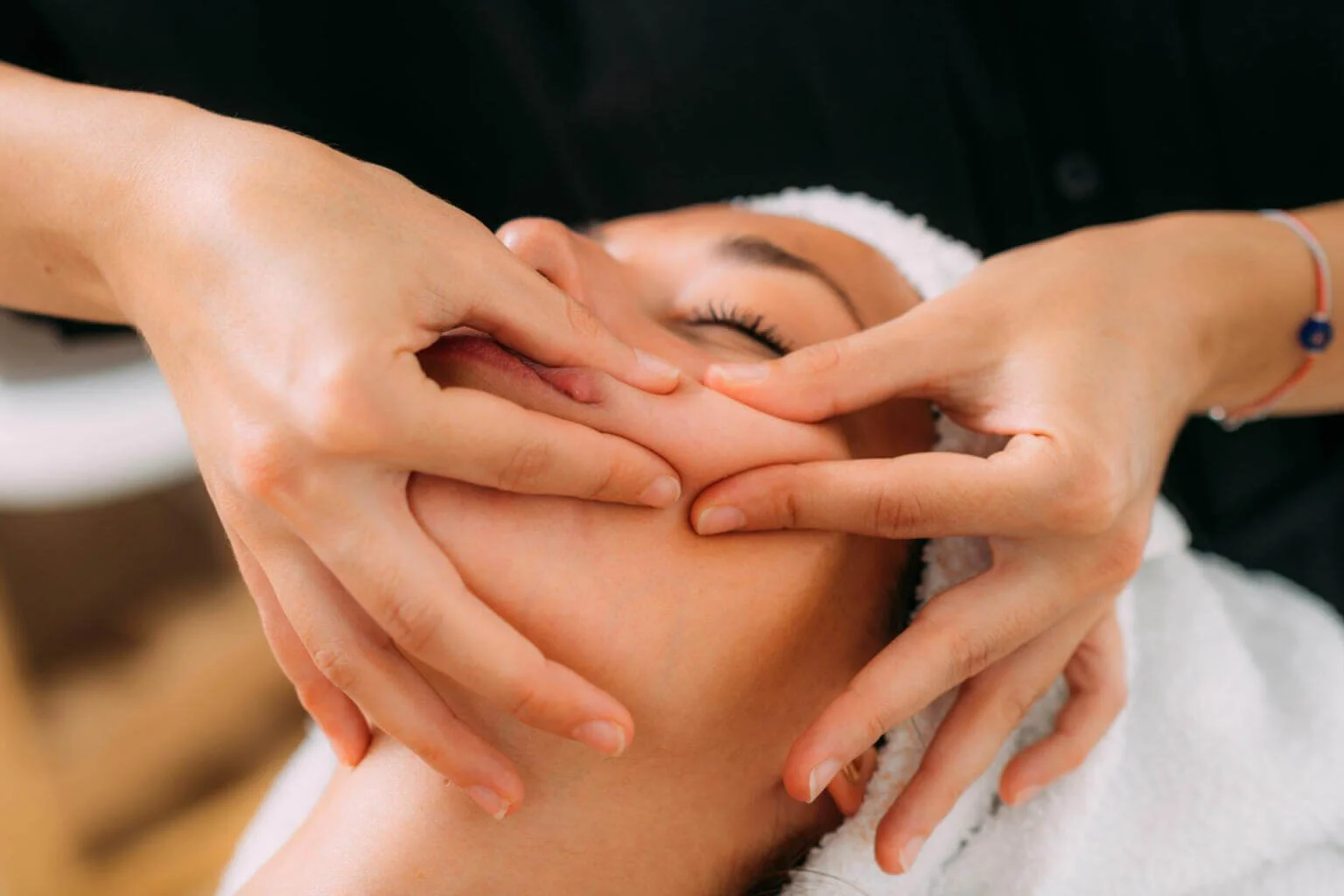Person receiving a facial massage with hands gently pressing on the face, eyes closed, wearing a white towel on head.