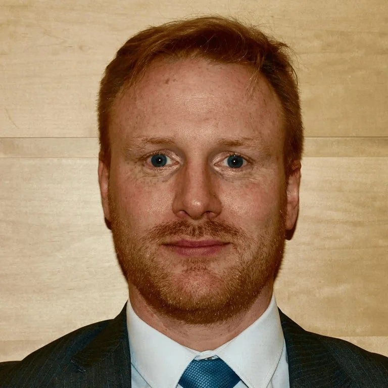 A man with red hair and a beard, wearing a suit and tie, standing in front of a light-colored wall.
