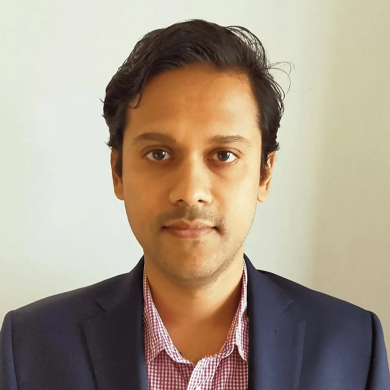 A man with dark hair wearing a dark suit and a red checkered shirt looking directly at the camera