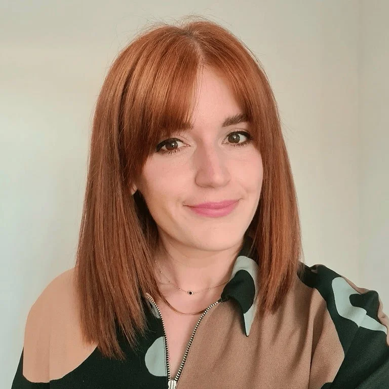 A woman with shoulder-length red hair and light skin, smiling, wearing a tan and black patterned top, in front of a plain light-colored wall.
