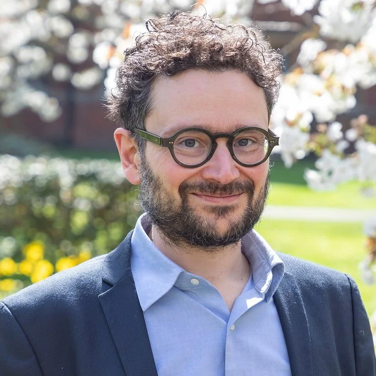 A man with curly hair and glasses smiling outdoors in front of blooming white flowers and greenery during daytime.