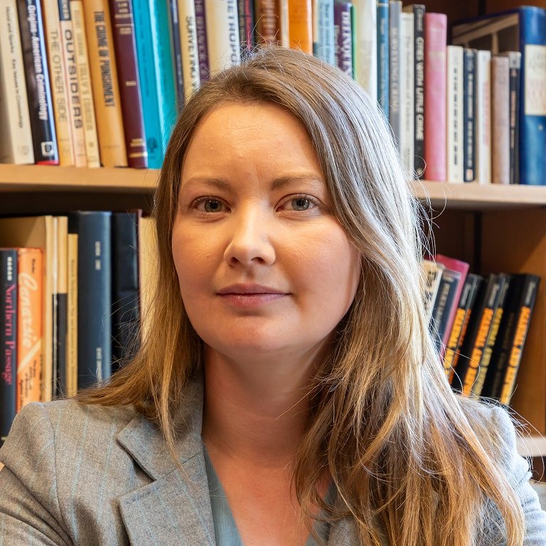 A woman with light brown hair, wearing a gray blazer, in front of a bookshelf filled with books.