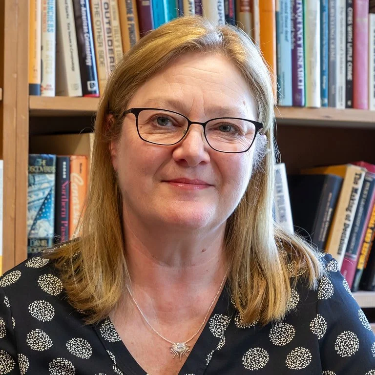 A woman with glasses and shoulder-length blonde hair, wearing a black dress with a white dotted pattern, smiling in front of a bookshelf filled with various books.