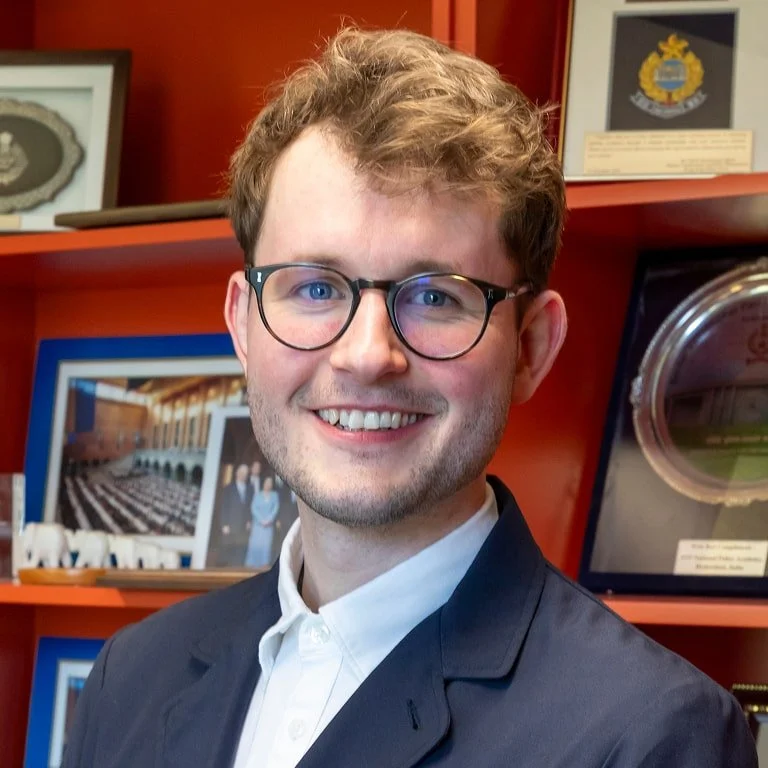 A man with glasses, light brown hair, and a light beard, smiling in front of a red background with framed photographs and awards.