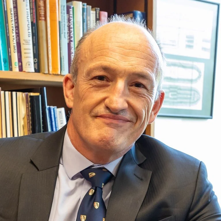 A middle-aged man in a suit and tie sitting in a library or office with bookshelves behind him and a framed document or certificate on the wall.