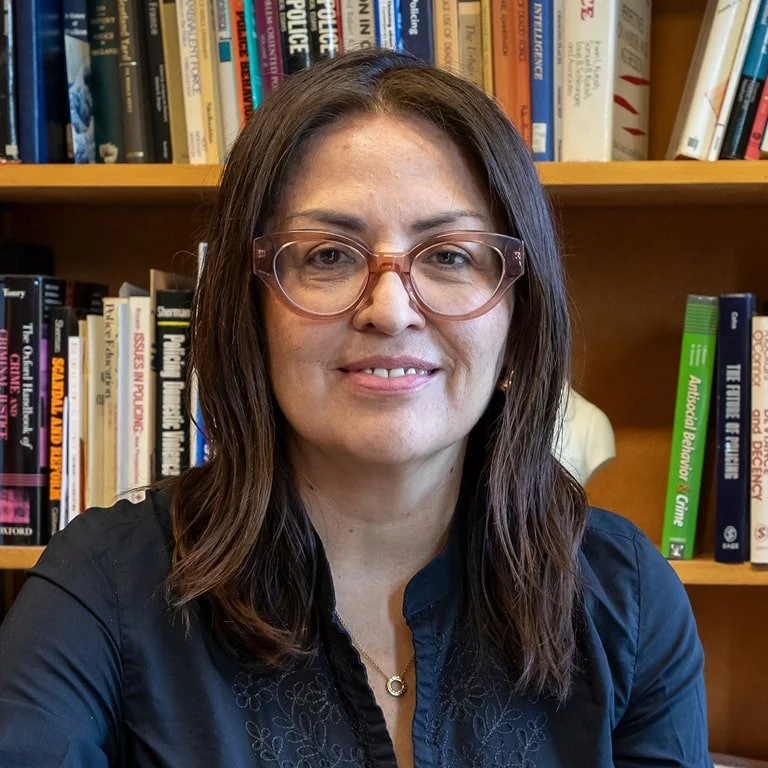 A woman with brown hair, wearing glasses, sitting in front of a bookshelf filled with books.