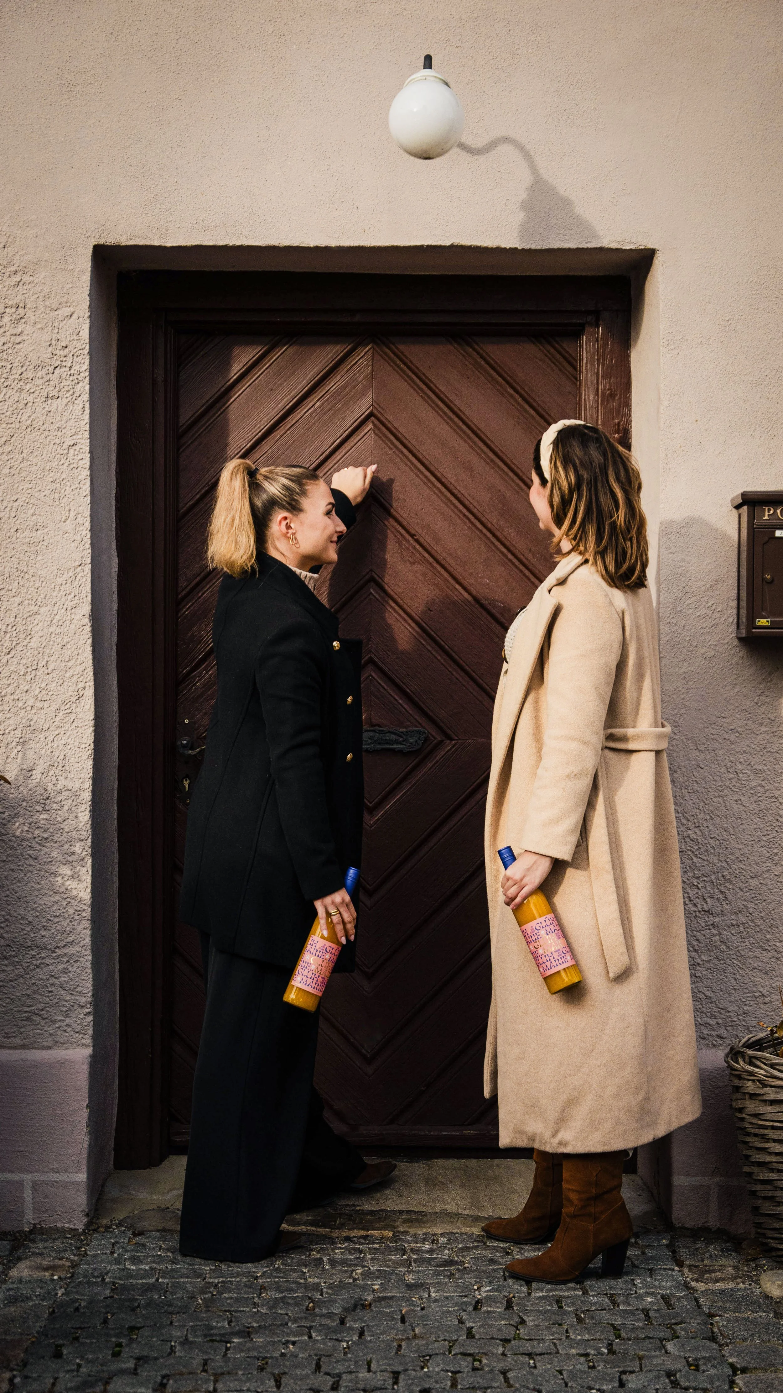 Zwei Frauen stehen vor einer Holztür und unterhalten sich, während sie Getränkeflaschen in den Händen halten.