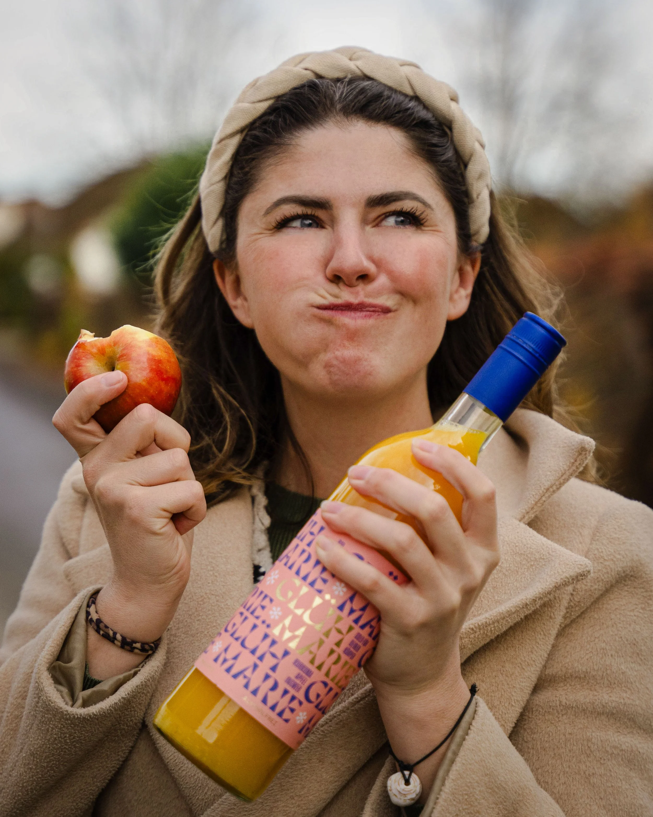 Eine junge Frau mit braunen Haaren, die einen Apfel in der linken Hand und eine Flasche mit Orangensaft in der rechten Hand hält, während sie draußen im Freien steht.