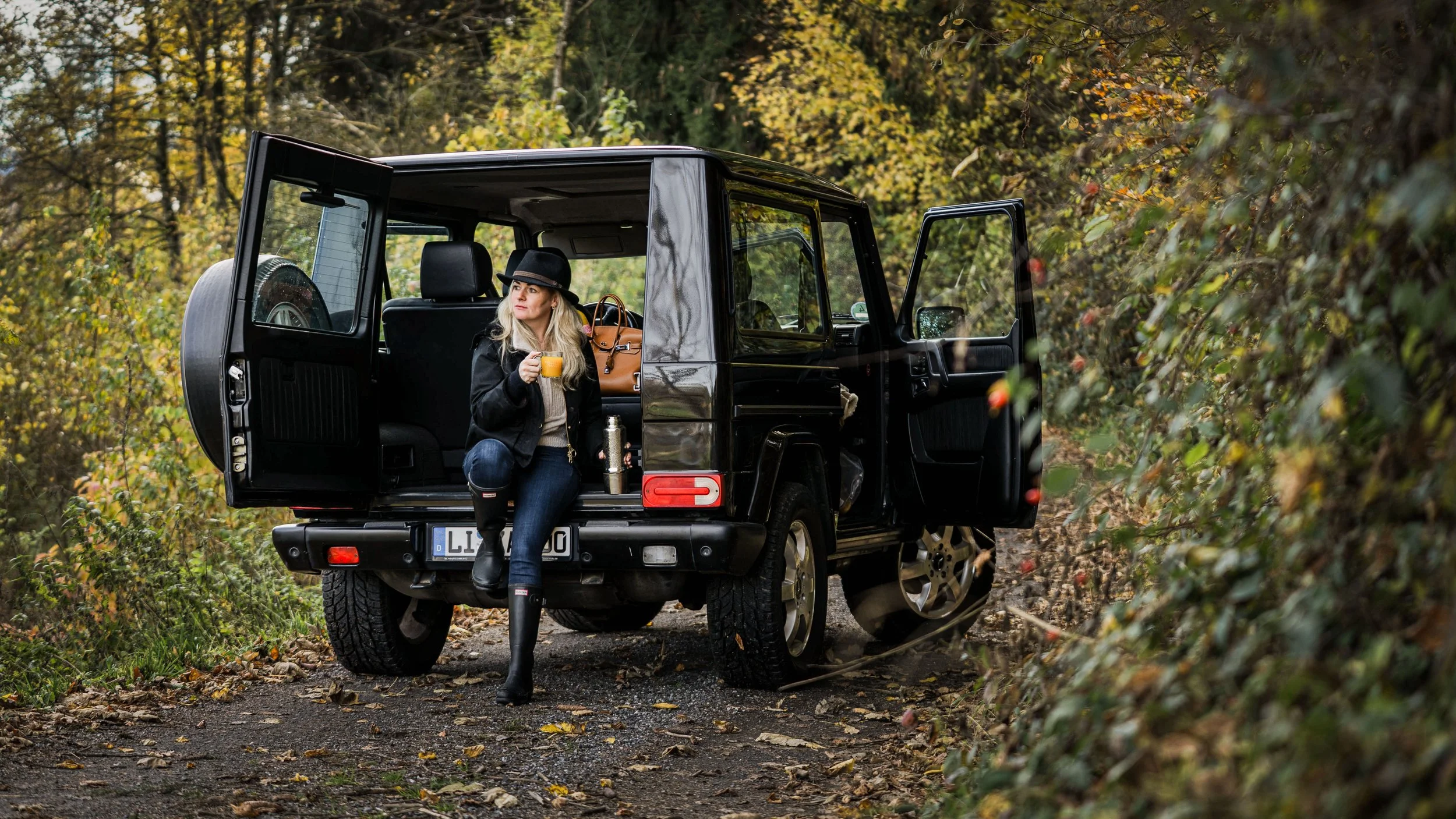Frau in Herbstkleidung mit Hut sitzt am Heck eines schwarzen Geländewagens, trinkt ein Glas Orangensaft, umgeben von Bäumen mit Herbstlaub.