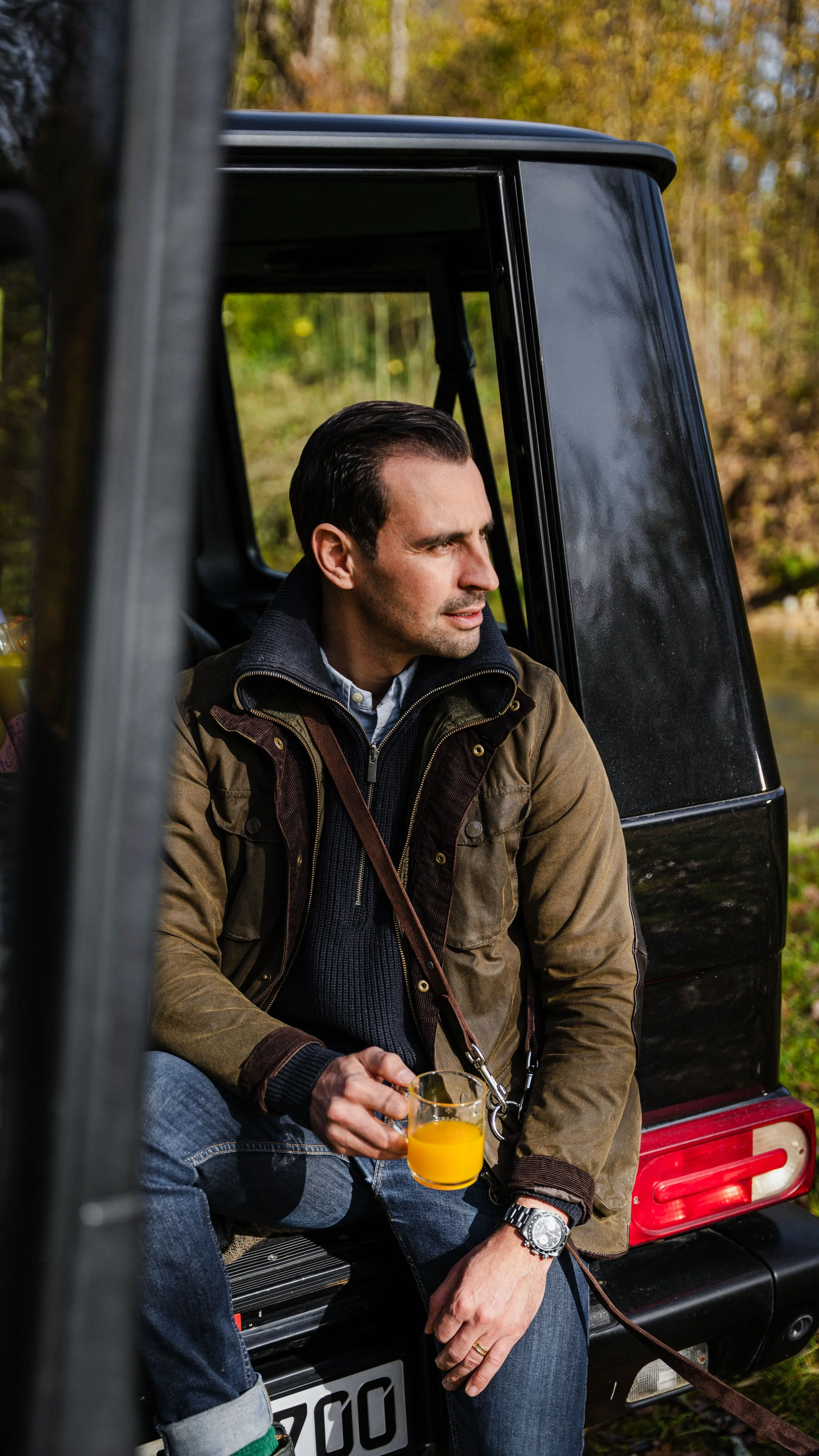 Mann sitzt auf der Stoßstange eines schwarzen Autos im Freien, hält ein Glas Orangensaft und schaut nachdenklich in die Ferne.