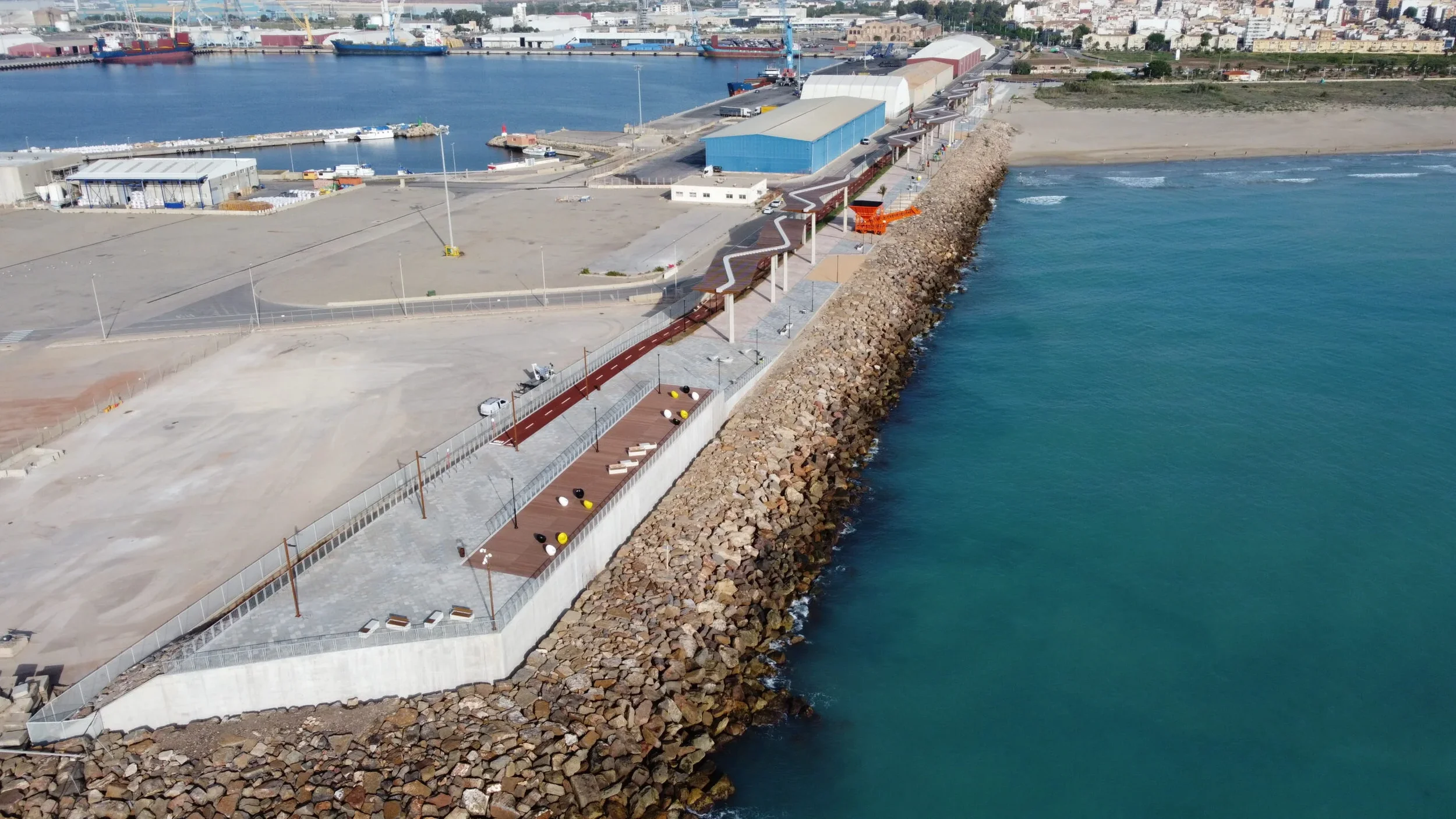 Vista aérea de un muelle y área portuaria en el mar con un rompeolas de rocas, varias estructuras de almacenamiento y almacenes en el fondo, y un camino peatonal con mobiliario en primer plano.