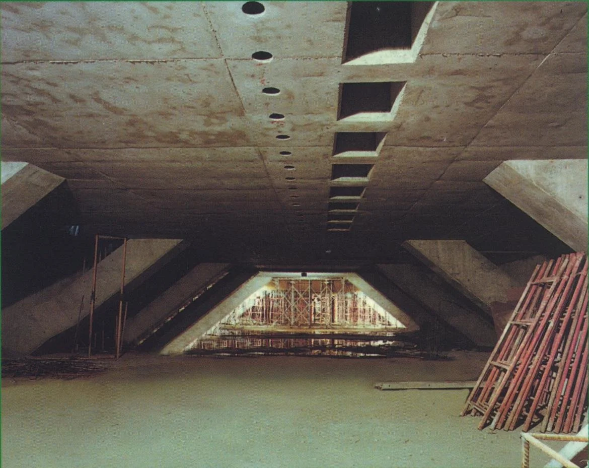 Vista del interior de un edificio en construcción, con un techo de concreto y aberturas cuadradas, con materiales de construcción y estructura de soporte en la parte inferior.