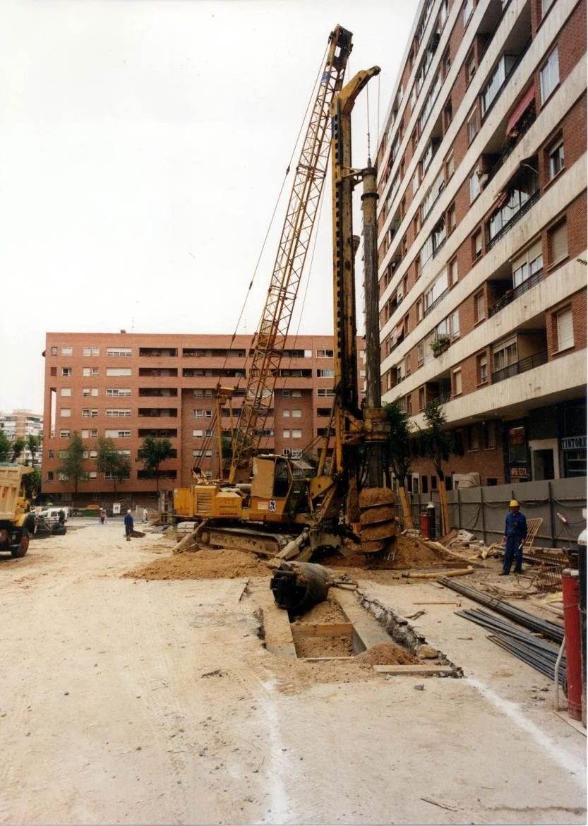 Maquinaria de construcción en una obra urbana, con edificios de apartamentos al fondo, trabajadores y materiales de construcción en el sitio.