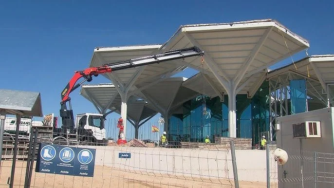 Construcción de una estación o edificio con techos modernos y estructura metálica, maquinaria de construcción en el sitio y trabajadores en labores de obra.