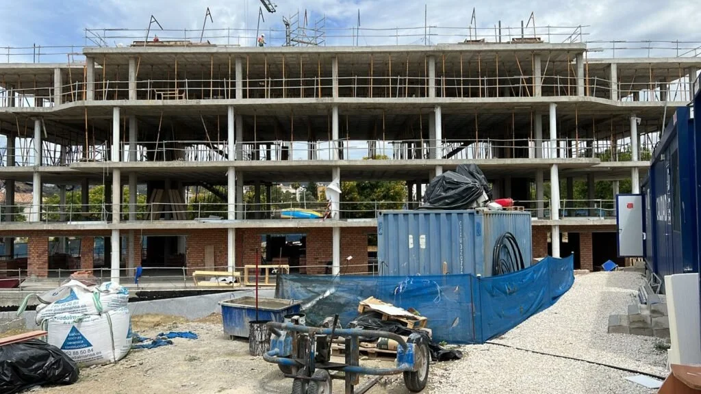 Construcción de un edificio de varias plantas con estructura de concreto y acero, en proceso de edificación. En primer plano, materiales y equipo de construcción, incluyendo sacos, contenedores y una estructura metálica. El sitio aún está en construc