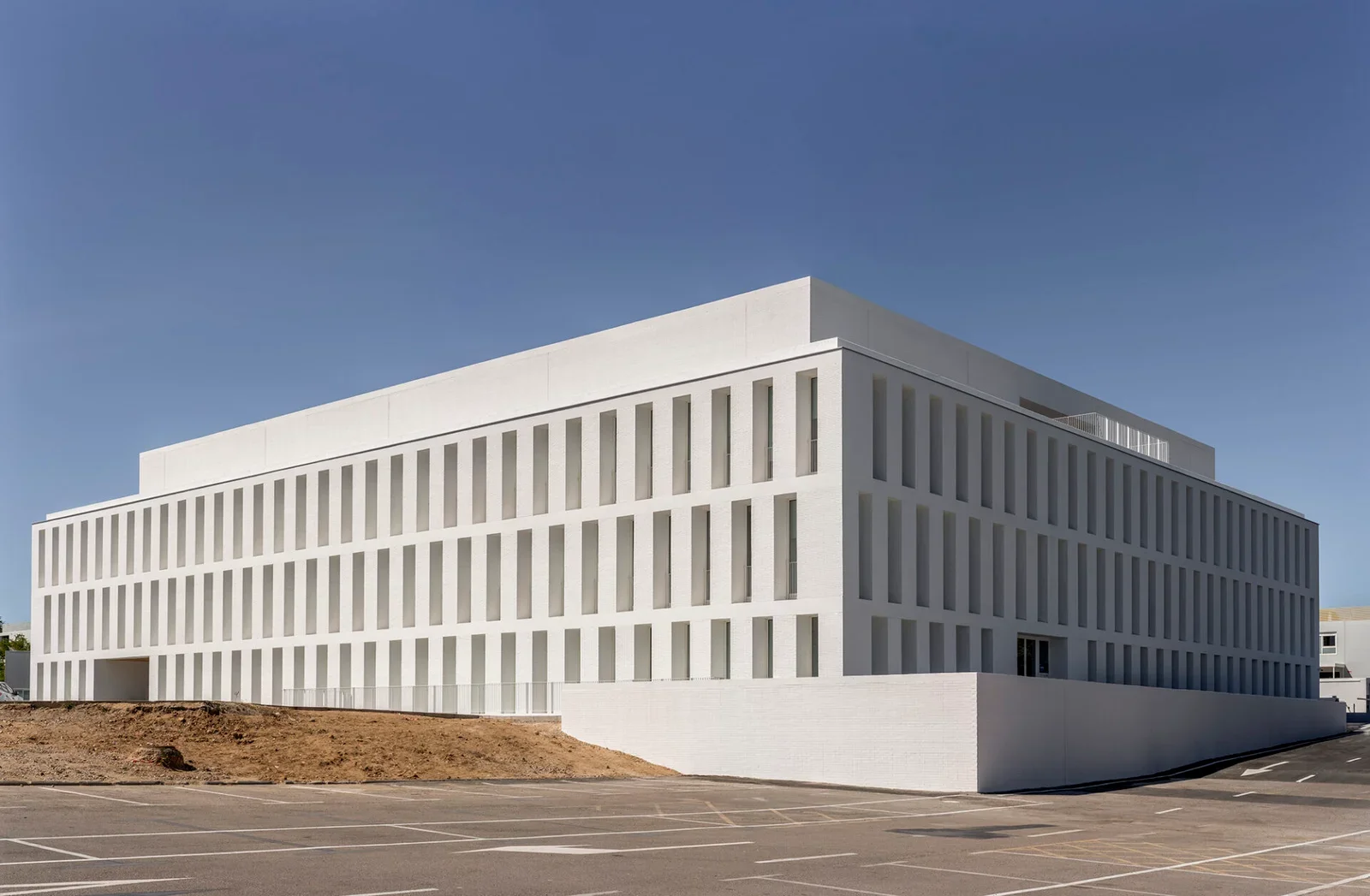 Edificio moderno de color blanco con ventanas altas y estrechas, en un entorno despejado con cielo azul y estacionamiento vacío en primer plano.