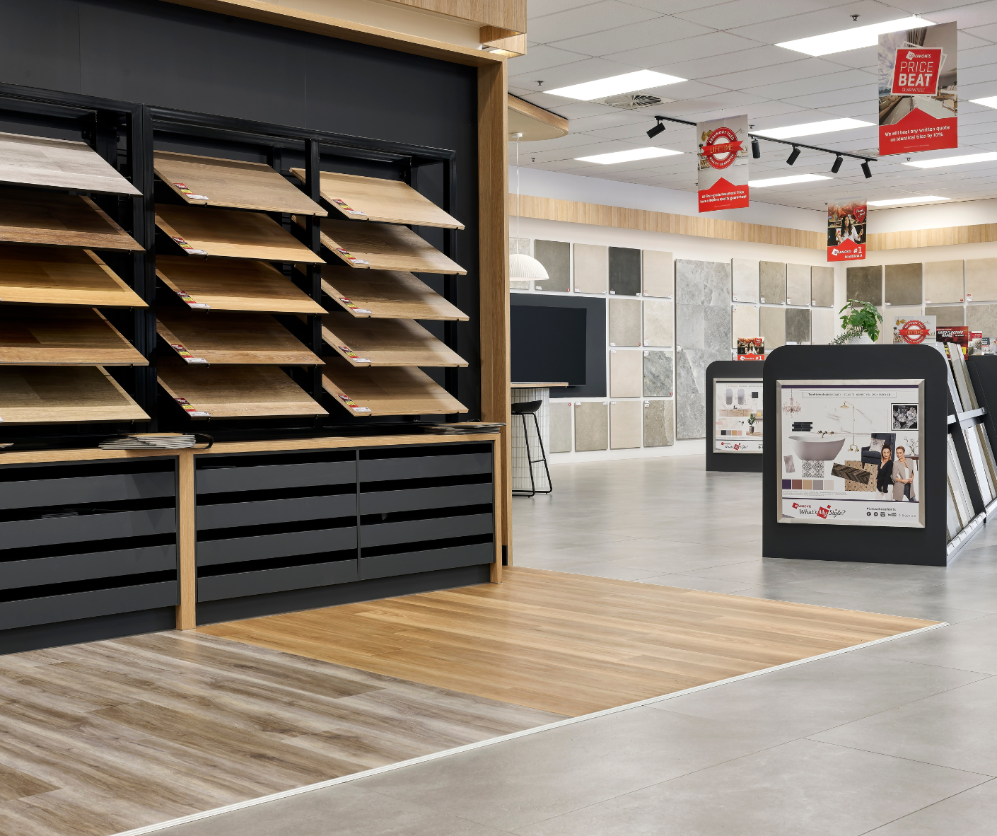 Showroom with display shelves of wood flooring samples, promotional signs hanging from ceiling, and sample tiles on a wall in the background.
