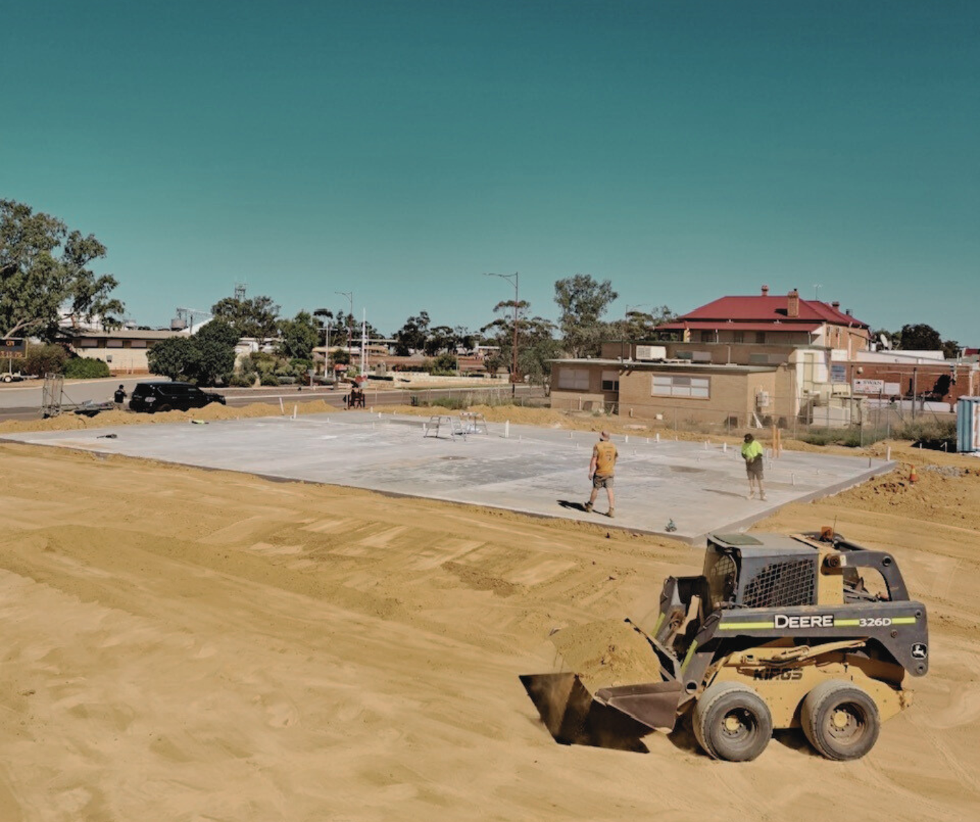 Construction site with a skid steer loader moving dirt in the foreground, and workers laying a concrete slab in the background with residential buildings and trees under a clear sky.