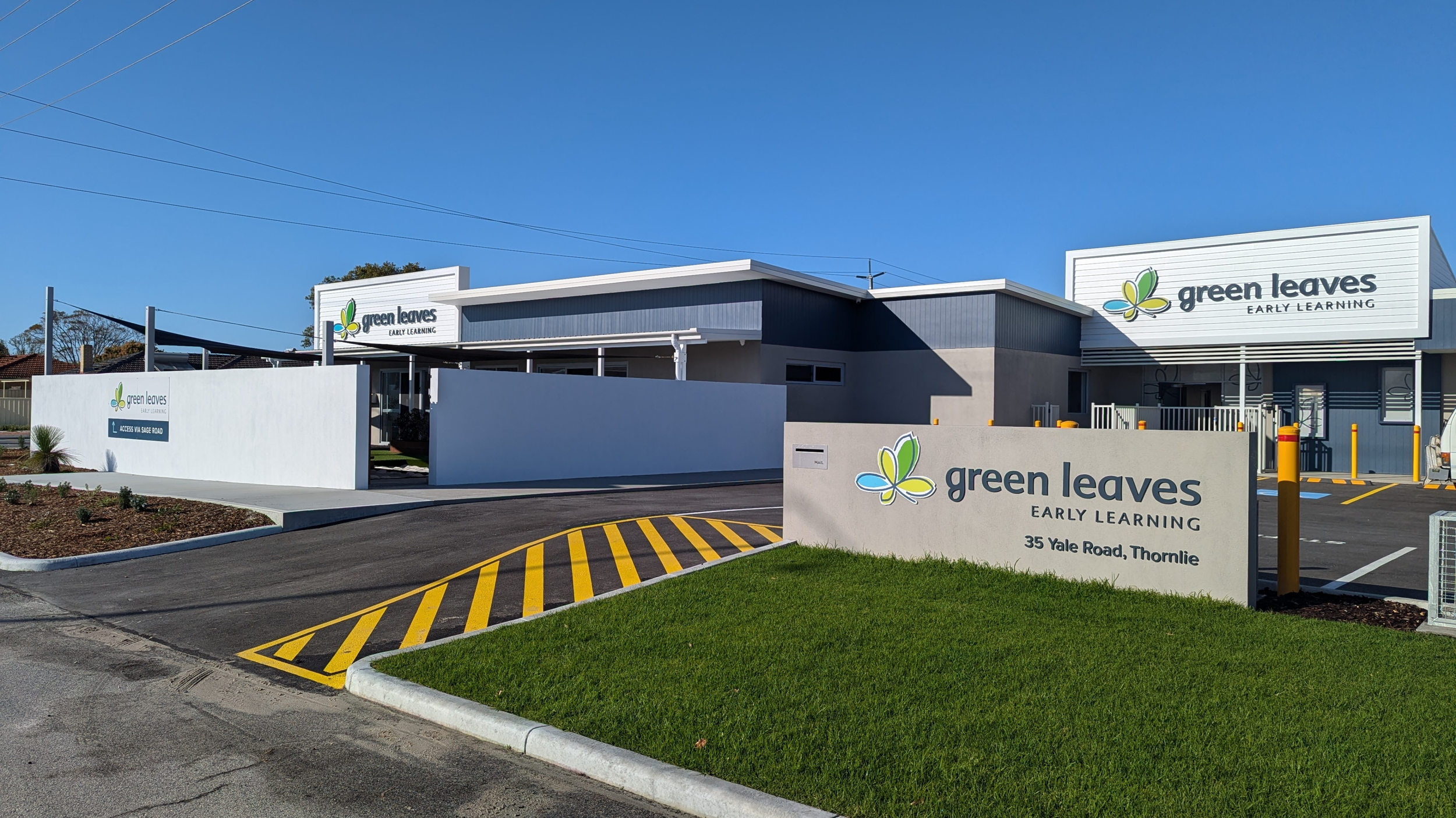 Exterior view of Green Leaves Early Learning preschool, with signs displaying its name and address at 35 Yale Road, Thornlie. The building features a modern design with blue and gray detailing, parking spaces, and well-maintained landscaping under a 