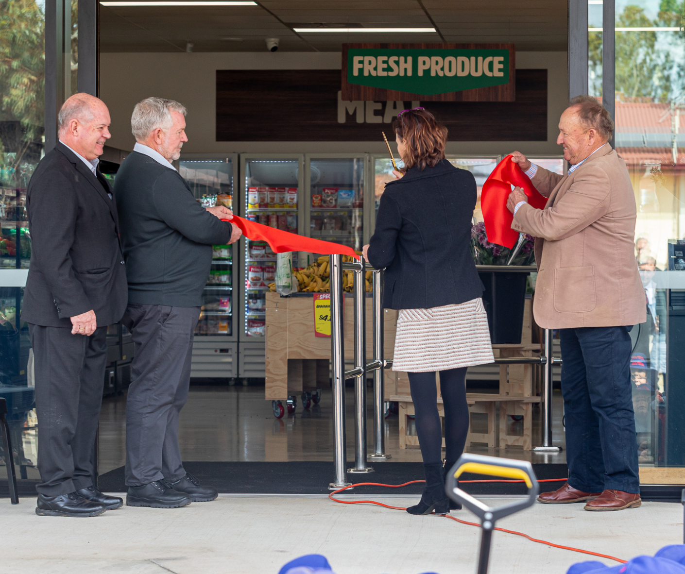 Four people participating in an opening ceremony for a fresh produce grocery store, with two men and a woman cutting a red ribbon, and a woman standing in front of them, inside the store showing fresh produce.