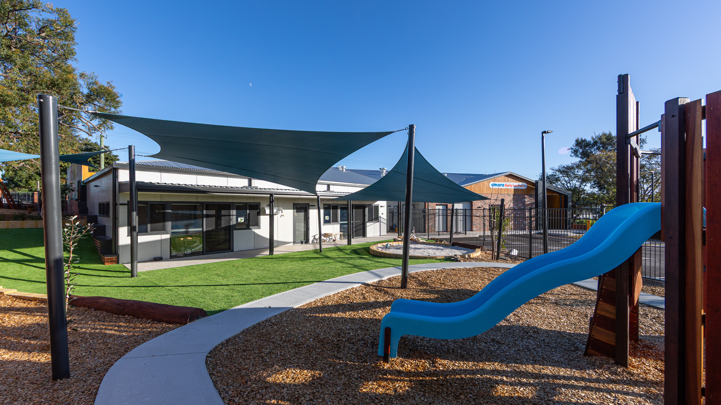 Playground with blue slide, shaded areas, and a building with a sign in the background on a sunny day.