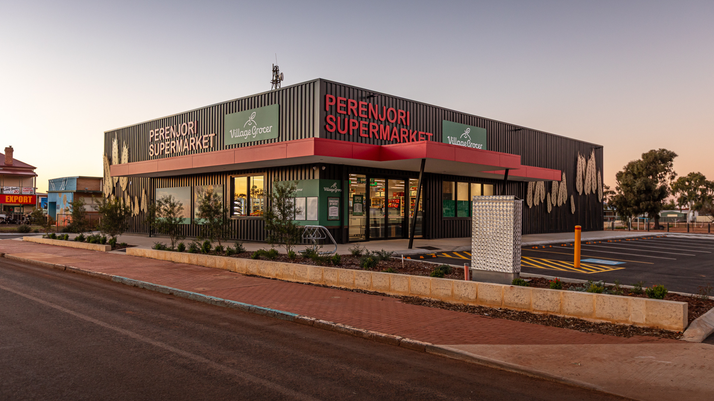 Exterior view of Perenjori Supermarket, a modern black building with red accents, large windows, and landscaping in front, during sunset.