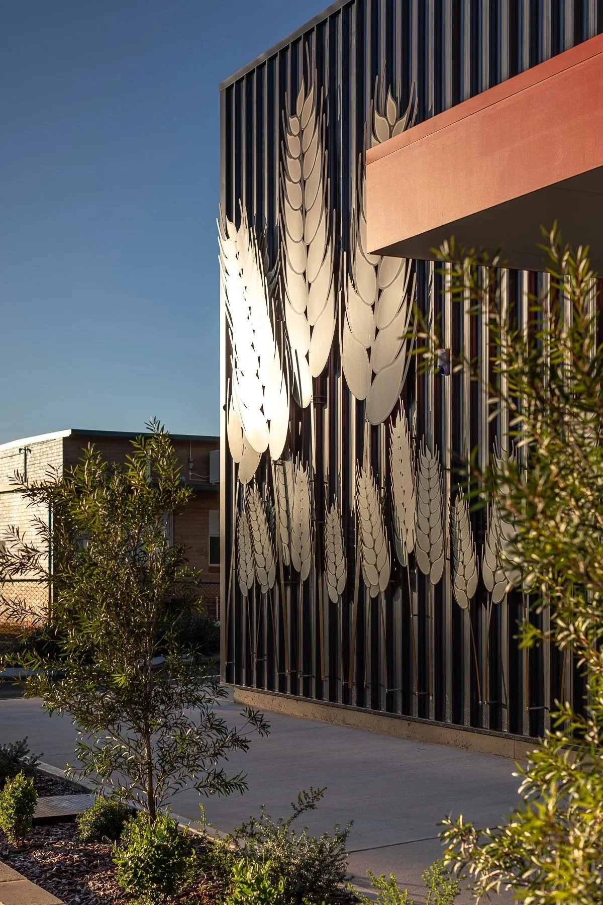 Modern building exterior with decorative metal panels that resemble leaves, with some trees and shrubs in the foreground and a clear blue sky.