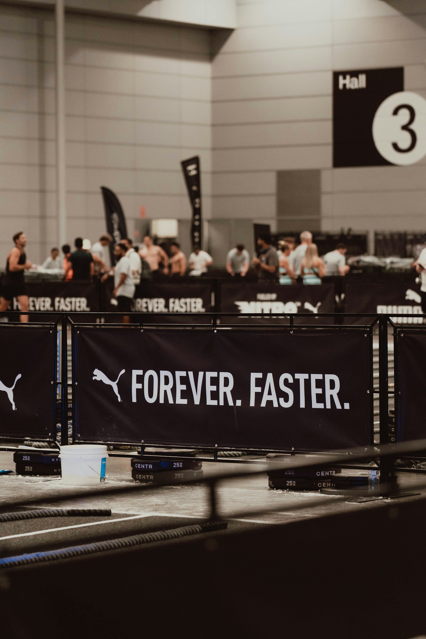 An indoor sports event with people gathering around a registration or check-in desk, and a large black banner with a white Puma logo and the slogan "FOREVER. FASTER." in the foreground.