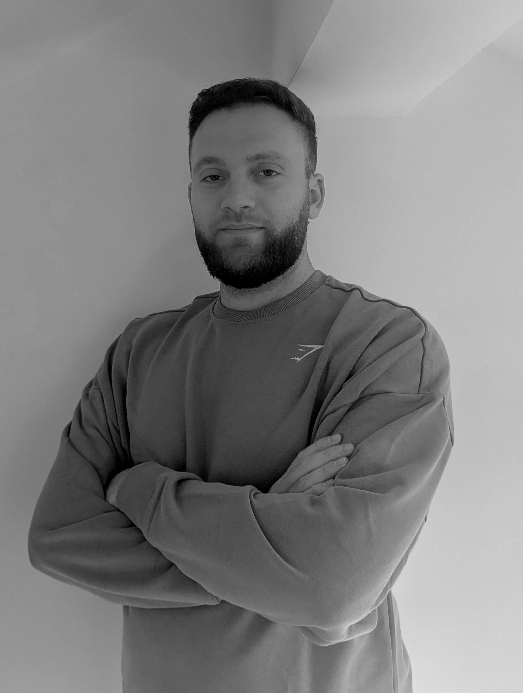 A man with a beard and short hair standing with folded arms against a plain wall, wearing a sweatshirt with a small logo on the chest.