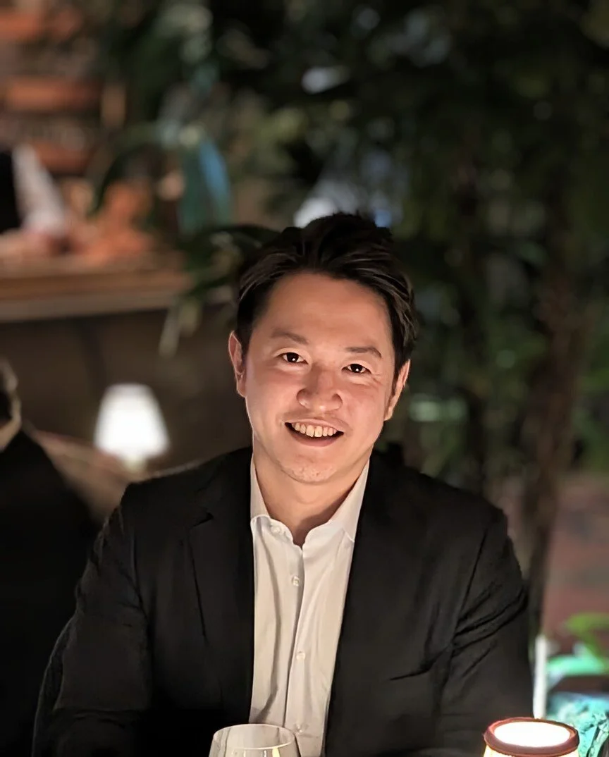 A young man with dark, styled hair wearing a white shirt and black blazer, smiling at the camera, sitting at a table in a dimly lit restaurant or bar with blurred greenery in the background.