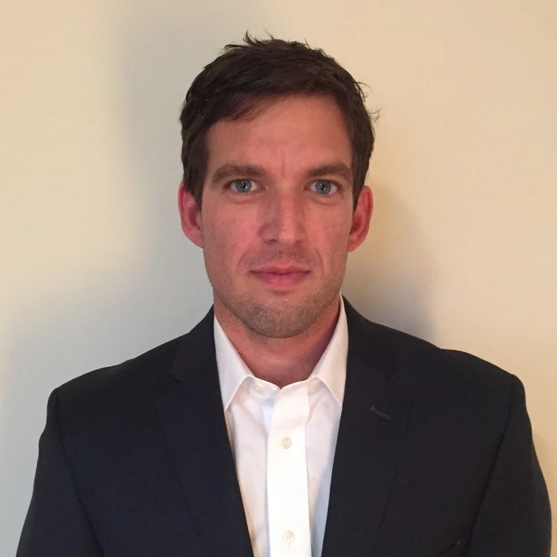 A man in a business suit with a white dress shirt, standing against a plain beige wall.