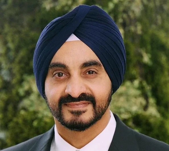 A man wearing a navy blue turban, black suit, and white shirt outdoors with green trees in the background.
