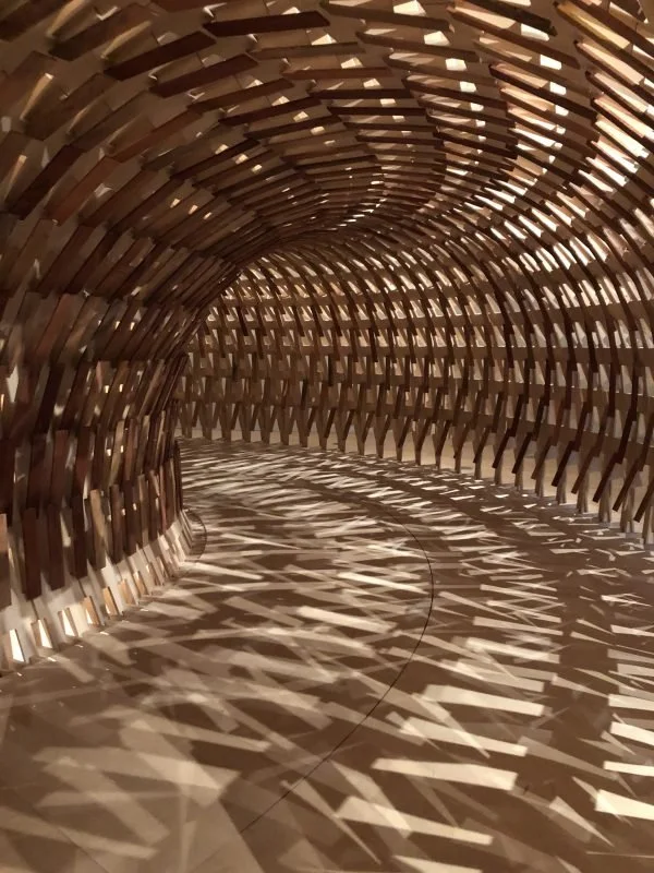 Interior view of a tunnel-like structure made of overlapping wooden slats, creating intricate shadow patterns on the floor.