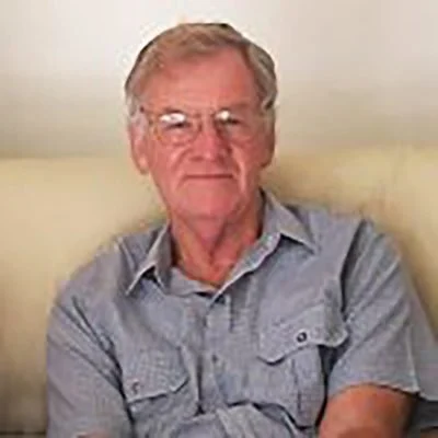 An elderly man with gray hair, glasses, and a light blue button-up shirt sitting against a plain wall.