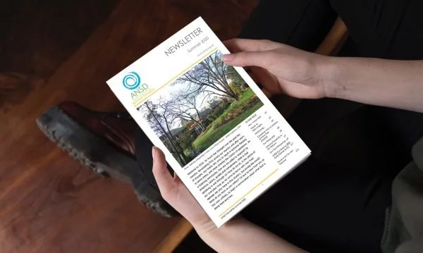 Person holding a newsletter with a photo of a park and trees on the front page, sitting next to a wooden table.