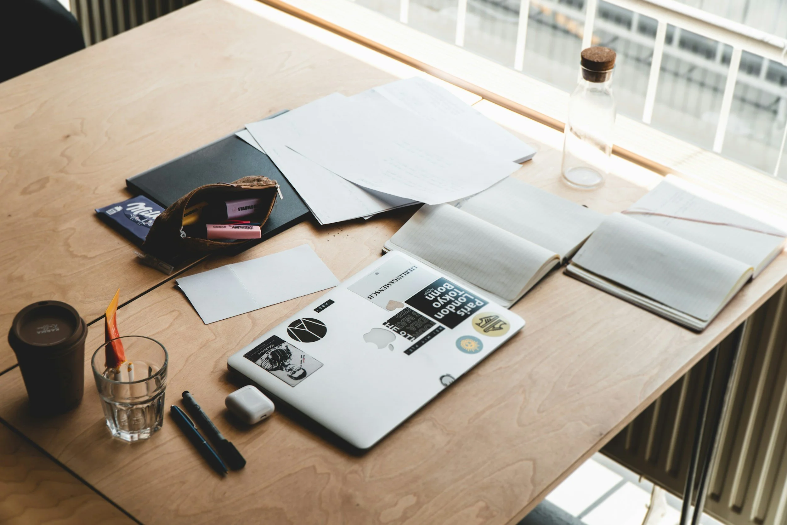 A wooden desk with scattered papers, a closed laptop with stickers, a black notebook, a pencil case, pens, a glass of water, a coffee cup, and a smartphone.– Olympus Migration | Andreas Athanasiou | Australian Visas