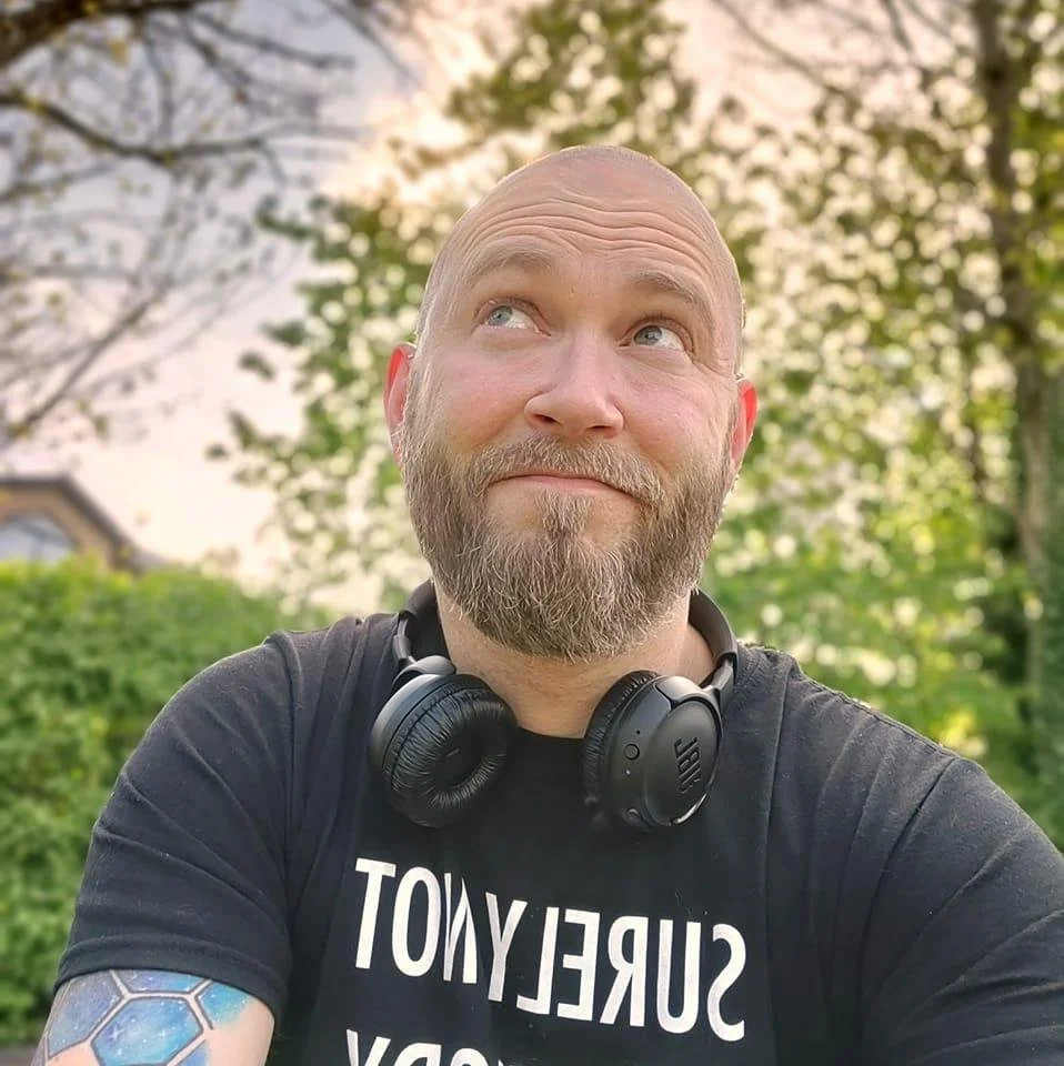 Mand med skæg, t-shirt med teksten 'SURREALITY', høretelefoner omkring halsen, stående udendørs med træer og blå himmel i baggrunden.