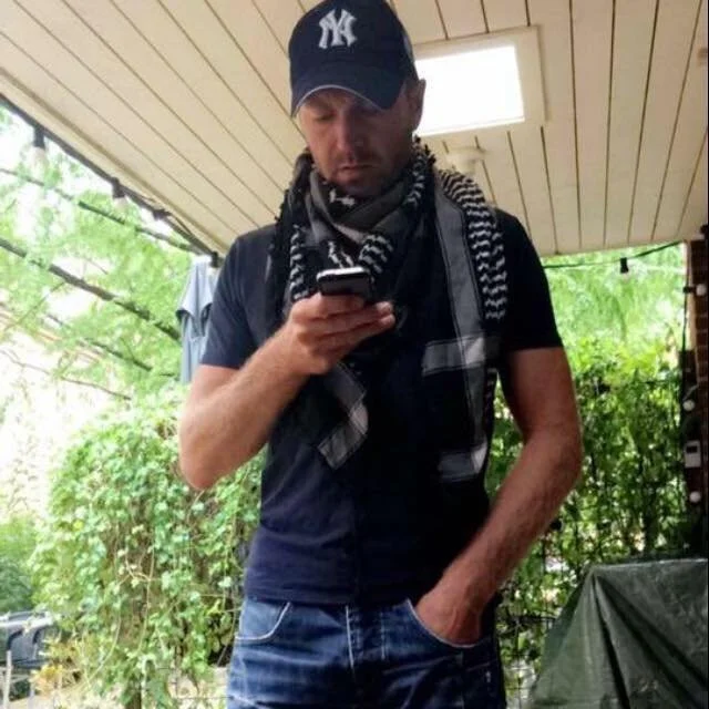 Mand iført en New York Yankees cap og sort t-shirt, der ser på sin telefon, med en sort og hvid stribet scarf om halsen, står under et træ eller en pergola, omgivet af grønne planter.