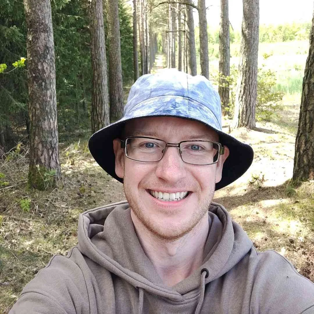 Mand tager selfie i en skov med træer og en sti bag ham.