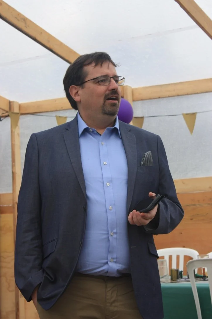 Mand i blazer og blå skjorte holder en smartphone i hånden, står indendørs med trævæge og pynt i baggrunden.