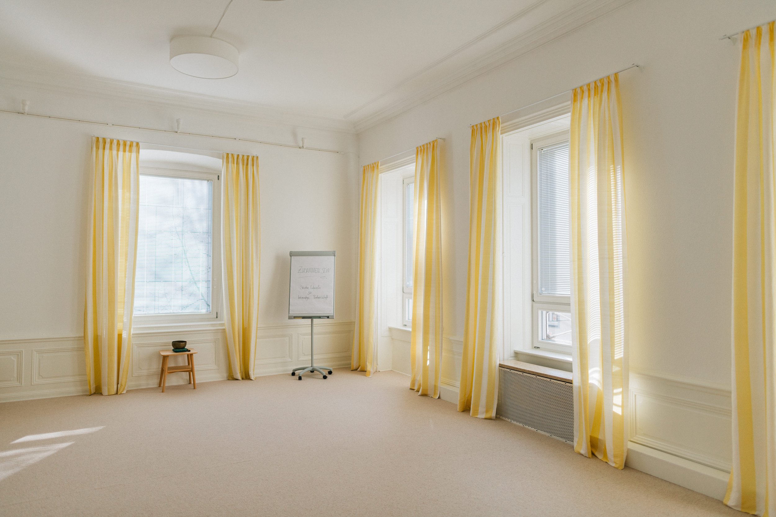 Ein heller Raum mit großen Fenstern, gelben Vorhängen und einer weißen Decke mit Stuckverzierungen. Es gibt einen kleinen Holzstuhl, einen Flipchart und eine Heizung unter den Fenstern.