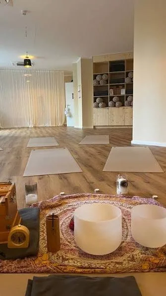 Yoga- oder Meditationsraum mit Yogamatten auf dem Boden, Klangschalen und anderen Meditationshilfsmitteln, in einem hellen Raum mit großen Fenstern und offenen Regalen mit Handtüchern oder Decken.