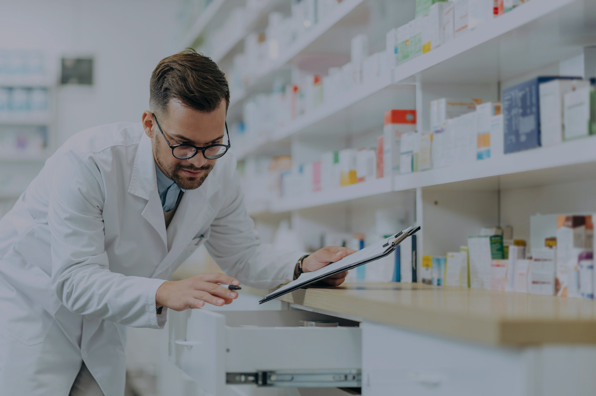 A male pharmacist with glasses wearing a white lab coat, holding a clipboard, and looking down at a drawer in a pharmacy aisle with shelves full of medicine boxes in the background.